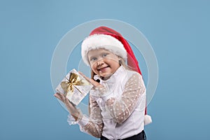 Waist up portrait of a little smiling blonde girl  with Sante Claus`s cap holding a gift box