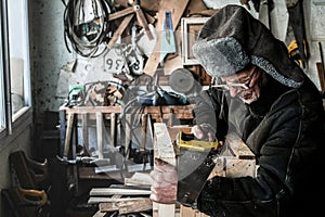 Waist up portrait of old grandfather in grey warm clothes in eyeglasses sawing wooden plank