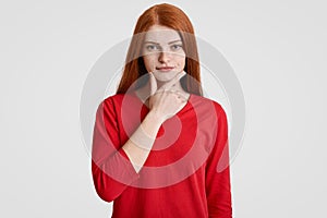 Waist up portrait of serious foxy female holds chin, has freckles skin, dressed in red casual sweater, being self confident, isola