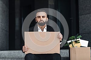 Waist up portrait of upset employee looking for job, sitting outside and holding empty poster, copy space