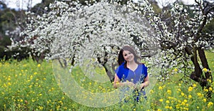 Waist up portrait of young woman in bright blue top in blossoming orchard and yellow mustard field, smiling, looking straight to t