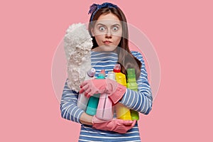 Waist up shot of angry housewife holds bottles of liquid detergent closely and brush, busy with housework, wears