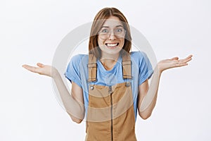 Waist-up shot of awkward cute charismatic young woman in brown overalls shrugging with hands near shoulders smiling with