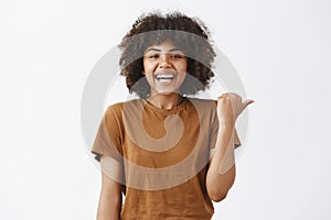 Waist-up shot of carefree joyful and attractive young dark-skinned female student in stylish t-shirt pointing right and