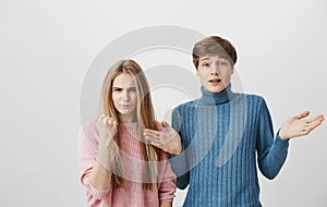 Waist-up shot of caucasian couple. Fair-haired male in blue sweater looks at camera with bewilderment, shrugs shoulders