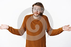 Waist-up shot of clueless unaware young bearded man in glasses with wavy hairstyle raising hands sideways and shrugging