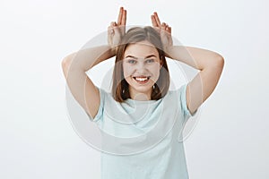 Waist-up shot of cute energized and playful emotive woman in trendy t-shirt holding fingers on head as if mimicking