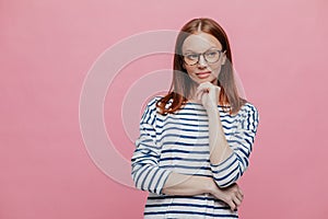 Waist up shot of dreamy thoughtful woman keeps hands partly crosed, holds chin, wears transparent glasses, looks pensively aside,