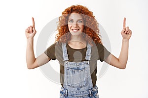 Waist-up shot friendly ambitious redhead curly woman show upper promo, pointing up to top, smiling delighted, introduce