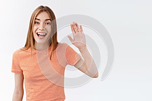 Waist-up shot friendly, pretty young woman in striped t-shirt, raising hand and waving as greeting you, lovely smiling