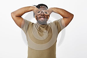 Waist-up shot of funny carefree african-american guy in olive t-shirt, holding hands over eyes and making faces