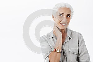 Waist-up shot of gentle delighted european senior female with white hair touching neck and smiling joyfully gazing at