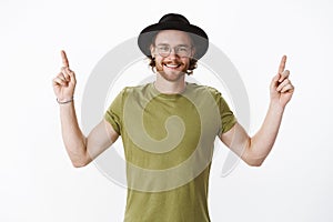 Waist-up shot of good-looking happy charismatic redhead bearded guy in hat and olive t-shirt raising hands pointing up