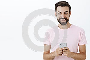 Waist-up shot of handsome delighted and carefree european man with beard and blue eyes holding smartphone looking