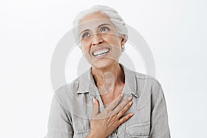 Waist-up shot of happy and delighted grateful charming old lady with white hair holding palm on chest in thankful