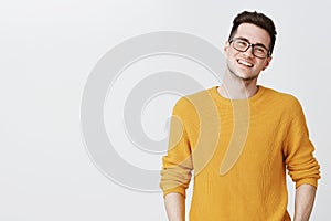 Waist-up shot of happy and delighted handsome young man in glasses and yellow sweater tilting head, smiling and laughing