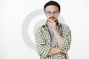 Waist-up shot of interested creative bearded young man in black beanie and glasses holding hand on chin and gazing with
