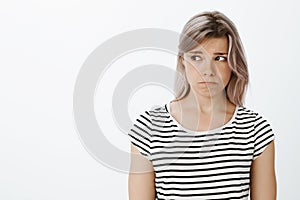 Waist-up shot of lonely gloomy cute girlfriend with fair hair in striped t-shirt, sulking and frowning, looking left