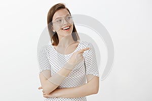 Waist-up shot of pleased and joyful good-looking woman in glasses, smiling broadly, gazing and pointing right with index