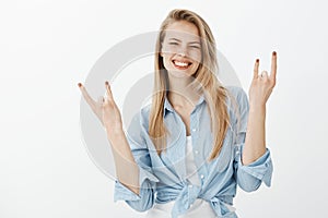 Waist-up shot of positive happy european blond woman in trendy clothes, raising hands and showing rock n roll sign