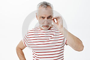 Waist-up shot of serious-looking suspicious old bearded man with grey hair looking from under glasses touching frames