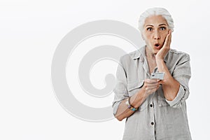 Waist-up shot of shocked and amazed grandmother exploring internet folding lips in amazement and surprise holding hand