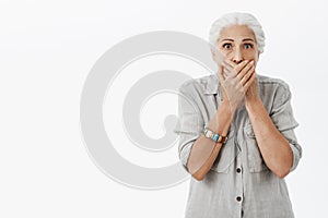 Waist-up shot of shocked and stunned elderly woman in casual shirt with grey hair gasping covering mouth with both palms