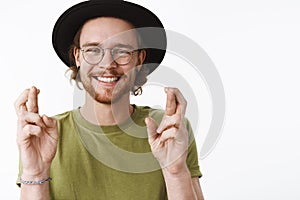 Waist-up shot of sincere happy and kind young bearded male traveler making hopes or wish with raised hands and crossed