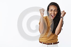 Waist-up shot of super excited astonished and happy joyful young african american woman raising clenched fists in