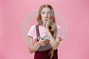 Waist-up shot of surprised amazed stunned young slender woman with wavy hair in overalls touching cheek from shock