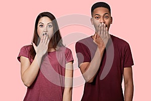 Waist up shot of surprised mixed race girlfriend and boyfriend being in stupor, cover mouth with palm, being shocked by terrific