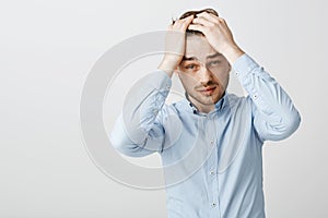 Waist-up shot of tired troubled good-looking young man with bristle pulling hair behind with hands on head frowning