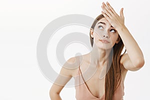 Waist-up shot of troubled beautiful caucasian female in beige evening dress holding hand on forehead and peeking up with