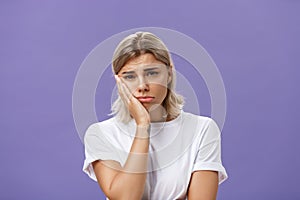 Waist-up shot of unhappy miserable and sad cute blonde female in white casual t-shirt pursing lips leaning face on palm