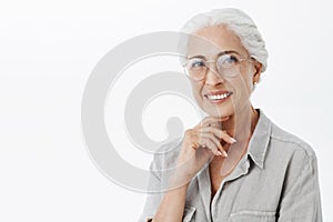 Waist-up shot of wise and pleased carefree kind grandmother in glasses with white hair holding hand on chin in