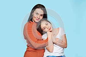 Waist up studio portrait of young mother embracing her daughter. Happy smiling family isolated over pastel blue background.