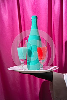 A waiter in black gloves holds a white tray with a turquoise and red wine glass and a bottle of champagne. Magenta