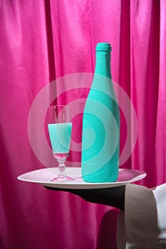 Waiter in black gloves holds a white tray with a turquoise wine glass and a bottle of champagne. Magenta curtains