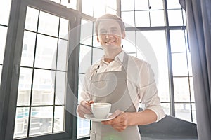 A waiter in a gray apron holds out a cup of coffee. Barista gives a cup of hot coffee in a cafe, against the background