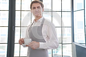 A waiter in a gray apron holds out a cup of coffee. Barista gives a cup of hot coffee in a café, against the background