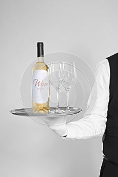 Waiter holding tray with glasses and bottle of wine on light background