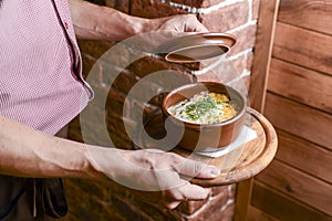 Waiter holds a bowl with potato gratin with cream and cheese and mushrooms in a restaurant. Waiter in a plaid uniform.