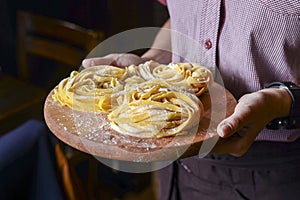 A waiter holds uncooked fettuccine nest with flour on wooden board. Tasty fresh ingredients for cooking pasta