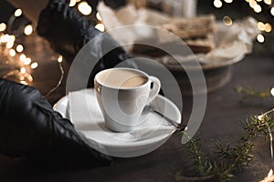 A waiter in medical gloves puts a cup of espresso on the table in coffee shop, Christmas bokeh lights. Selective focus