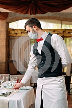 A waiter in a medical protective mask serves the table in the restaurant.