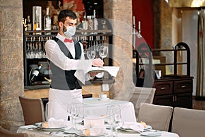 A waiter in a medical protective mask serves the table in the restaurant.