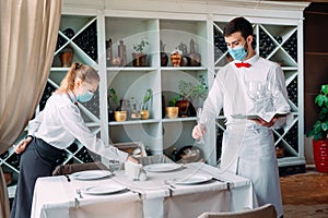 A waiter in a medical protective mask serves a table in the restaurant. Employees of a restaurant or hotel in protective