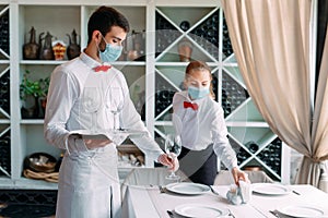A waiter in a medical protective mask serves a table in the restaurant. Employees of a restaurant or hotel in protective