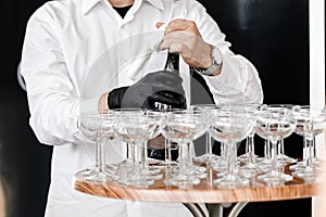 The waiter opens a bottle of champagne, sparkling wine. waiter in black gloves pours champagne into glasses on wooden