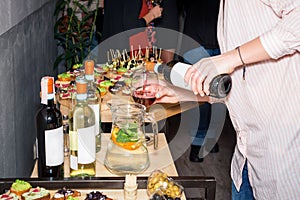 Waiter pouring red wine in a glass at a restaurant table full of appetizers with guests standing near.